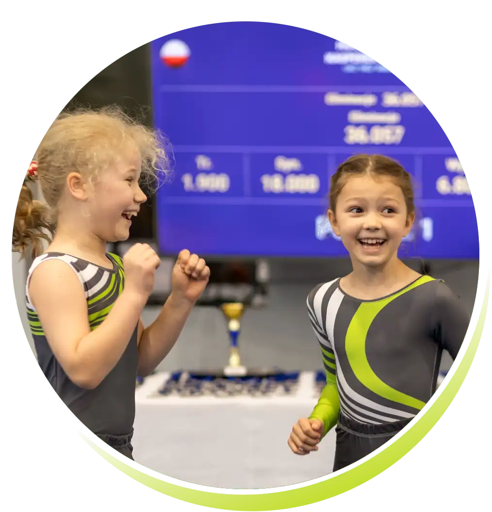 Dwie dziewczynki w klubowych strojach skaczą z radości, w tle puchar i wyniki zawodów z biało-czerwoną flagą